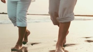 Gün batımında sahilde çıplak ayakla yürüyen romantik bir çiftin kırpılmış görüntüleri.