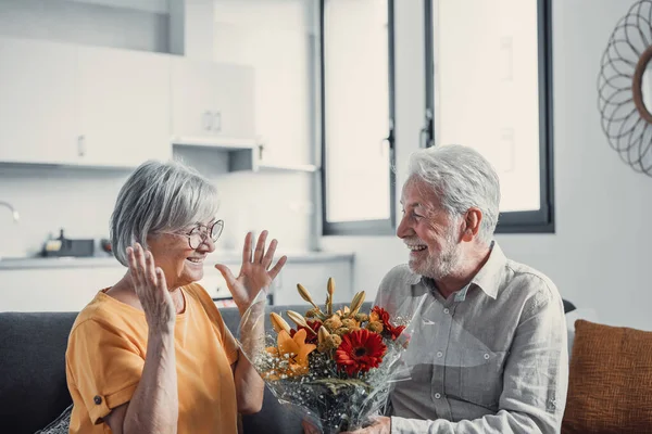 stock image Old man giving flowers at his wife sitting on the sofa at home for the San Valentines day. Pensioners enjoying surprise together. In love people having fun. 