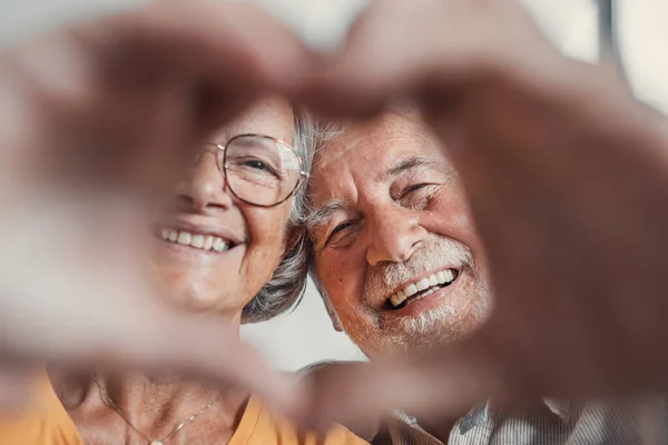 Stock image Close up portrait happy sincere middle aged elderly retired family couple making heart gesture with fingers, showing love or demonstrating sincere feelings together indoors, looking at camera.