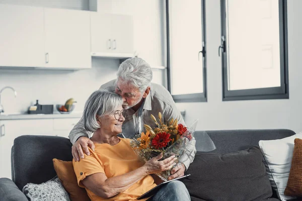 Old man giving flowers at his wife sitting on the sofa at home for the San Valentines day. Pensioners enjoying surprise together. In love people having fun.