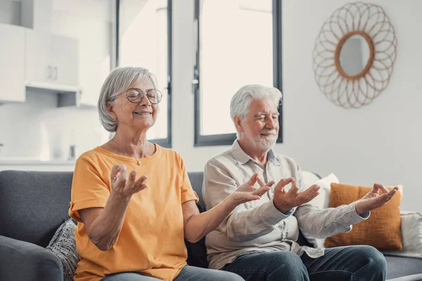 Calm senior middle aged couple practicing yoga together sitting in lotus pose on sofa, mindful peaceful mature man and woman meditating relaxing in living room at home, old people healthy lifestyle