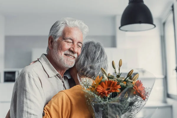 Old man giving flowers at his wife sitting on the sofa at home for the San Valentines day. Pensioners enjoying surprise together. In love people having fun.