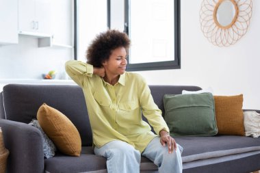 Üzgün Afrikalı genç bir kadın masajdan yorgun düşmüş, boynu tutulmuş, evdeki kanepede izole edilmiş, yorgun, yorgun, siyah bir kadın omuz ağrısını hafifletmek için kaslarını ovuyor, fibromiyalji konsepti.