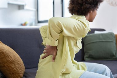 Afro öğrenci kız, evdeki oturma odasında kanepede otururken sırt ağrısı çekiyor. Bilgisayarda uzun süre çalıştıktan sonra sırt ağrısı çekiyor. Sabit çalışma ve kötü duruş konsepti