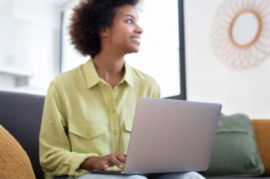 Gülümseyen genç siyahi kadın dizüstü bilgisayar kullanıyor, rahat bir koltukta dinleniyor, video görüşmesinde konuşuyor, internette sohbet ediyor, internette geziniyor, film izliyor, internetten alışveriş yapıyor. Tembel dijital cihaz kullanıcısı