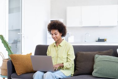 Gülümseyen genç siyahi kadın dizüstü bilgisayar kullanıyor, rahat bir koltukta dinleniyor, video görüşmesinde konuşuyor, internette sohbet ediyor, internette geziniyor, film izliyor, internetten alışveriş yapıyor. Tembel dijital cihaz kullanıcısı