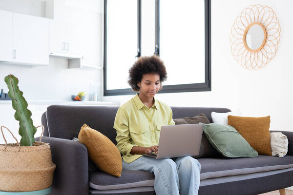 Smiling young Black woman using laptop computer, resting on comfortable couch, talking on video call, chatting online, browsing internet, watching movie, shopping on internet. Lazy digital gadget user