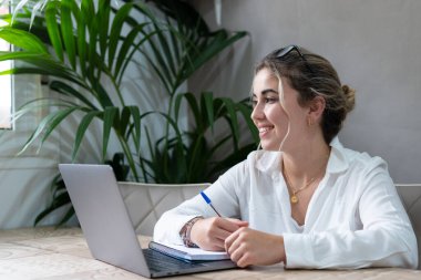 Gülümseyen bin yıllık beyaz kız öğrencinin portresi evde bilgisayarla çalışırken masada oturuyor. Mutlu, genç, neşeli bir kadın bilgisayar kullanıyor. Uzak bir kursa ya da eğitime gidiyor. Eğitim kavramı.