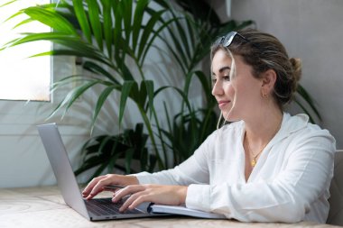 Milenyum tebessüm eden beyaz kız, oturma odasında oturmuş dizüstü bilgisayarla notlar alıyor, bilgisayarla uğraşıyor, deftere yazıyor, evde online kurs veya eğitim görüyor, eğitim kavramı geliştiriyor.