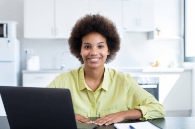 Neşeli, kısa saçlı, iş yerinde laptopuyla oturan, kameraya bakan, gülümseyen kısa saçlı siyahi bir iş kadını. Milenyum çalışanı, çalışanı, girişimci portresi