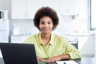 Neşeli, kısa saçlı, iş yerinde laptopuyla oturan, kameraya bakan, gülümseyen kısa saçlı siyahi bir iş kadını. Milenyum çalışanı, çalışanı, girişimci portresi