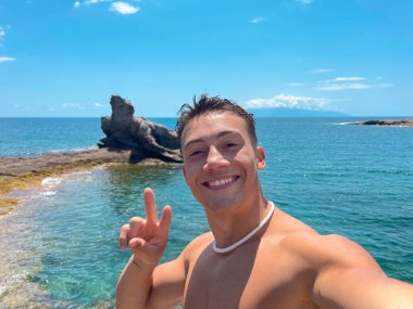 Sahilde gülümseyen, kameraya bakıp telefonuyla selfie çeken çekici ve mutlu bir genç adam. Yaz tatilinde eğleniyor ve eğleniyoruz.