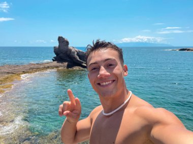 Sahilde gülümseyen, kameraya bakıp telefonuyla selfie çeken çekici ve mutlu bir genç adam. Yaz tatilinde eğleniyor ve eğleniyoruz.