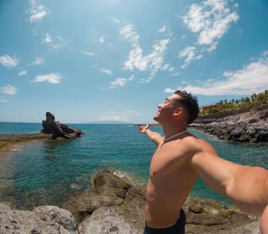 Sahilde gülümseyen ve okyanusa bakan çekici ve mutlu bir genç adam. Kamerayı tutuyor ve telefonuyla selfie çekiyor. Yaz tatilinde eğleniyor ve eğleniyoruz.