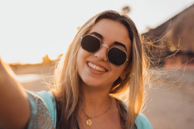 Genç, güzel, sarışın bir kızın, elinde fotoğraf makinesiyle sahilde fotoğraf çektirip arka planda günbatımının keyfini çıkarırken çekilmiş bir portre..