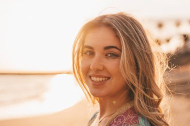 Mutlu, gülümseyen, kumsalda poz veren sarışın kadın, günbatımı gökyüzü ve arka planda bulanık deniz. 