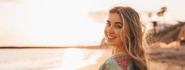 Mutlu, gülümseyen, kumsalda poz veren sarışın kadın, günbatımı gökyüzü ve arka planda bulanık deniz. 