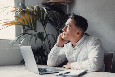 Düşünceli işadamı çenesine dokunuyor, fikirleri ya da stratejileri düşünüyor, dizüstü bilgisayarla ahşap çalışma masasında oturuyor. 