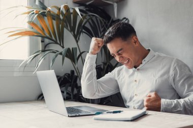 Dizüstü bilgisayarı olan mutlu bir adam başarılı, kazananın mutlu duyguları, arka planda ev bitkisi  