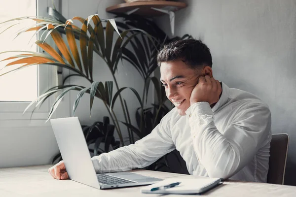 stock image handsome young businessman with laptop at desk remotely, Pleasant happy man communicating in social network, house plant on background 