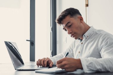 Side view handsome young businessman in eyewear working with computer remotely, sitting at wooden table in office. Pleasant happy man communicating in social network, searching information online.