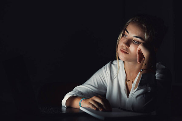 Уставшая молодая женщина сидит за рабочим столом после окончания рабочего дня в одиночестве в массажных храмах, пытаясь вспомнить важную информацию. Уставшая женщина-работница чувствует головную боль, вызванную работой сверхурочно