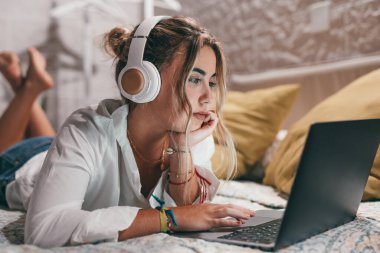 Evde dizüstü bilgisayar kullanan, kulaklık takmış ve müzik dinleyen genç ve güzel bir kadını yakından çek. Milenyum gençleri çevrimiçi çalışırken eğleniyor..