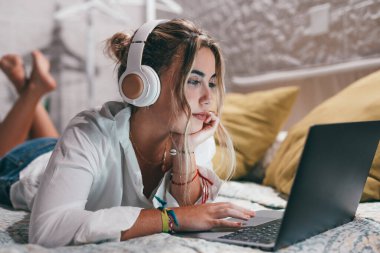 Evde dizüstü bilgisayar kullanan, kulaklık takıp müzik dinleyen genç ve mutlu bir kadını yakından çek. Milenyum gençleri çevrimiçi çalışırken eğleniyor..