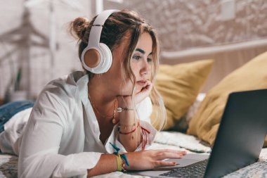 Evde dizüstü bilgisayar kullanan, kulaklık takmış ve müzik dinleyen genç ve güzel bir kadını yakından çek. Milenyum gençleri çevrimiçi çalışırken eğleniyor..