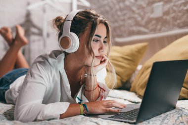 Evde dizüstü bilgisayar kullanan, kulaklık takıp müzik dinleyen genç ve mutlu bir kadını yakından çek. Milenyum gençleri çevrimiçi çalışırken eğleniyor..