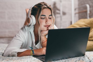 Evde dizüstü bilgisayar kullanan, kulaklık takıp müzik dinleyen genç ve mutlu bir kadını yakından çek. Milenyum gençleri gözleri kapalı şarkıların tadını çıkarıyor.