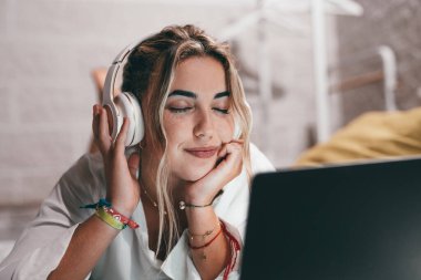 Evde dizüstü bilgisayar kullanan, kulaklık takıp müzik dinleyen genç ve mutlu bir kadını yakından çek. Milenyum gençleri gözleri kapalı şarkıların tadını çıkarıyor.