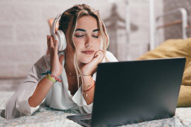 Evde dizüstü bilgisayar kullanan, kulaklık takıp müzik dinleyen genç ve mutlu bir kadını yakından çek. Milenyum gençleri gözleri kapalı şarkıların tadını çıkarıyor.