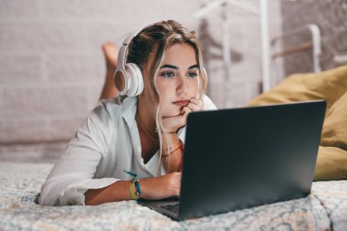 Evde dizüstü bilgisayar kullanan, kulaklık takmış ve müzik dinleyen genç ve güzel bir kadını yakından çek. Milenyum gençleri çevrimiçi çalışırken eğleniyor..