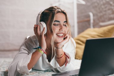 Evde dizüstü bilgisayar kullanan, kulaklık takıp müzik dinleyen genç ve mutlu bir kadını yakından çek. Milenyum gençleri gözleri kapalı şarkıların tadını çıkarıyor.