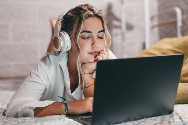 Evde dizüstü bilgisayar kullanan, kulaklık takıp müzik dinleyen genç ve mutlu bir kadını yakından çek. Milenyum gençleri gözleri kapalı şarkıların tadını çıkarıyor.