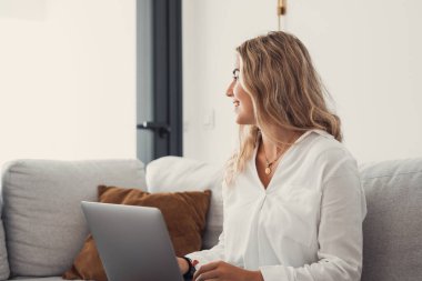 Çekici sarışın bir kadının portresi dizüstü bilgisayarını kanepenin üzerinde kullanıyor evde internette geziniyor ve pencereye bakıyor.