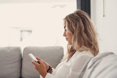 Çekici, sarışın bir kadının portresi. Kanepede cep telefonu kullanıyor. Evde internette geziniyor. Çevrimiçi konsept yaşam tarzı rahatlıyor