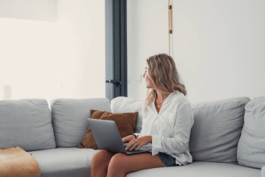 Çekici sarışın bir kadının portresi dizüstü bilgisayarını kanepenin üzerinde kullanıyor evde internette geziniyor ve pencereye bakıyor.