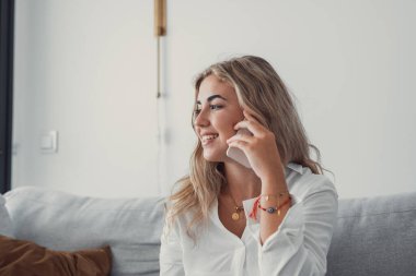 Mutlu, neşeli genç bir kadın evde telefonla konuşuyor, gülümseyen genç bir kız cep telefonuyla arıyor koltukta oturuyor, güzel bir bayan cep telefonuyla eğlenceli bir konuşma yapıyor.