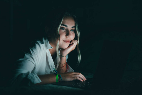 Улыбается уверенный в себе студент, использующий ноутбук ночью в постели, работающий, заканчивающий проект в поздние часы, дедлайн, смотрящий на экран компьютера, сидящий в спальне для домашней работы в университете