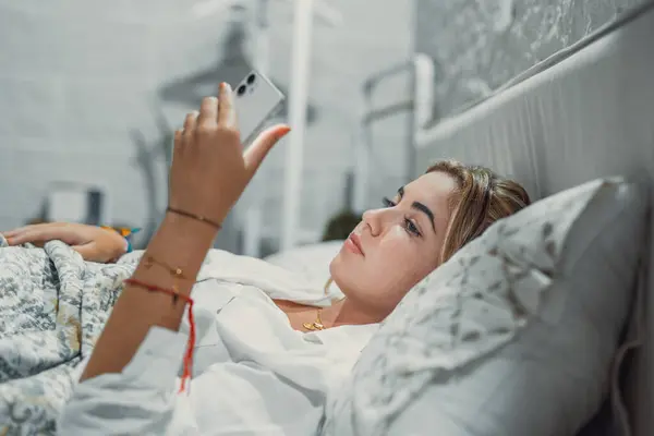 Gadget addiction. Calm bored young woman or teenage girl lying at cozy bed holding cellphone in hands looking at screen chatting checking social network account before fall asleep or after waking up
