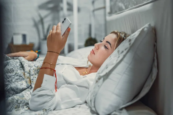 Gadget addiction. Calm bored young woman or teenage girl lying at cozy bed holding cellphone in hands looking at screen chatting checking social network account before fall asleep or after waking up