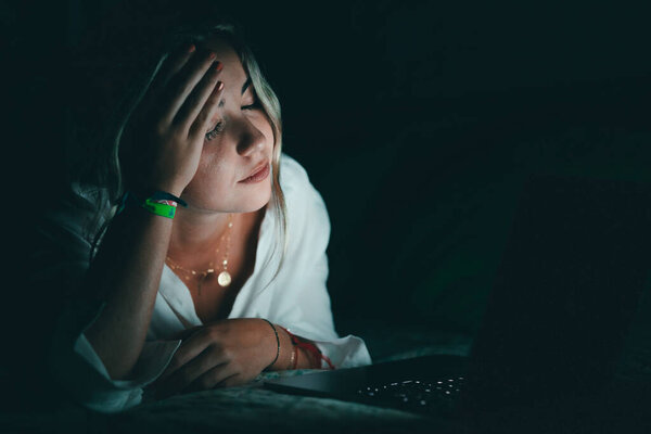 Ошеломленный юный кавказский студент касается головы, страдает от расплывчатого зрения, массирует носовой мост, снимает боль, чувствует себя уставшим, работая допоздна за компьютером в постели.