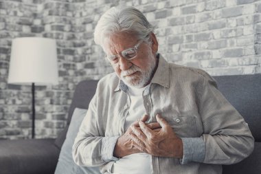 Worried elder senior man feeling bad, upset old middle aged grandfather touching chest feel sudden pain heartburn having heart attack sit on sofa at home