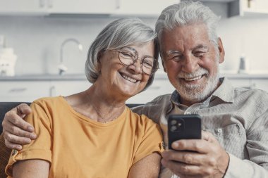 Mutlu olgun aile, eş ve koca birlikte evde telefon kullanarak, kanepede oturan orta yaşlı erkek ve kadın gülümseyen, mobil cihaz uygulamaları kullanarak, sosyal ağda video izlerken, internette sörf
