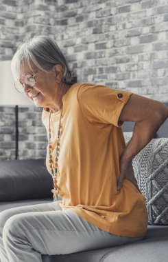 Unhappy mature woman touching back, feeling pain, sitting on couch in living room, unhealthy middle aged senior female suffering from backache, spinal problem, rubbing stiff muscles or kidney clipart