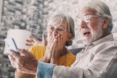 Happy older couple sit on couch staring at cellphone screen gesturing looking overjoyed, scream with joy, read fantastic news, get great commercial offer, pension raise. Success, achievement concept