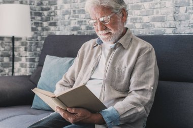 Headshot portrait close up of old happy and relaxed man sitting reading a book at home. Mature male person enjoying free time having fun indoor.