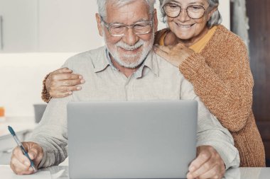 Yaşlılık ve modern teknoloji. Emekli aile çifti mutfak masasında oturup internetten son haberleri izleyen dizüstü bilgisayarlı fotoğraf videosu kullanıyor. Yaşlı koca paylaşımı, üst düzey eşle web sitesinde ilginç bilgiler hakkında tartışır